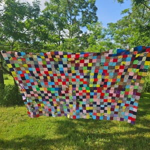 Op de afbeelding: Een grote rechthoekige quilt opgebouwd uit een raster van talloze kleine stoffen vierkantjes in een grote verscheidenheid aan effen kleuren en diverse patronen, waaronder strepen, stippen en bloemenprints, horizontaal hangend aan een waslijn.