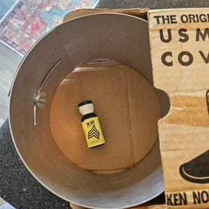 May include: A cylindrical metal container with a wing nut fastener on the side, containing a small bottle with a yellow label featuring the text 'M-NU' and a chevron graphic. A piece of cardboard with the text 'THE ORIGINAL USM COVER' is visible.