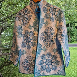 May include: A quilted jacket featuring a light cream base with a repeating teal and dark blue floral medallion pattern. The garment has a high collar, long sleeves with cuffs, and vertical stitching throughout, finished with teal piping along the edges.