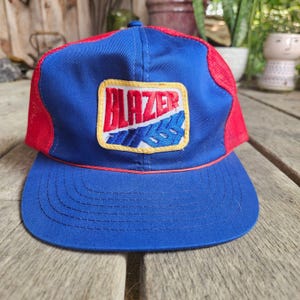 May include: A blue and red trucker cap featuring a rectangular patch on the front panel. The patch displays the word BLAZER in bold red letters above a blue graphic design, all outlined in yellow. The cap has a solid blue brim and mesh back.