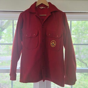 Puede incluir: Una camisa de lana roja de manga larga con cuello clásico, cierre frontal de botones y dos bolsillos en el pecho con solapas abotonadas. Un parche circular amarillo y rojo con las letras BSA y un emblema de águila está cosido en el bolsillo.