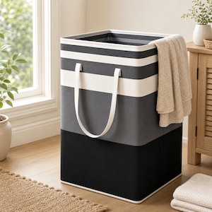 May include: A square laundry hamper featuring horizontal stripes in black, dark grey, and white. The hamper has two white fabric handles attached to the front and a beige towel draped over the top edge, creating a functional storage container.