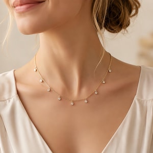 May include: A gold chain necklace featuring multiple small, round, clear stones spaced evenly along the length of the chain. The necklace rests against the neck, showcasing a delicate design with individual bezel-set stones hanging from the links.