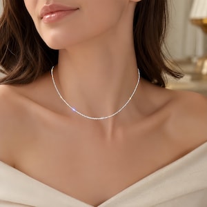 May include: A thin, shimmering silver-toned chain necklace that rests closely around the base of the neck. The chain features a textured, reflective surface that catches the light, creating a subtle sparkle along the entire length of the metal strand.