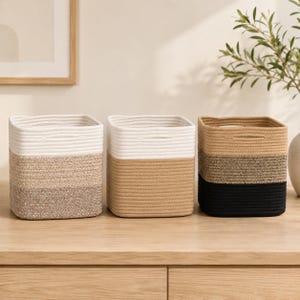 May include: Three square storage baskets made of coiled rope, each featuring integrated side handles. The first basket has white and tan sections, the second is white and light brown, and the third is tan, dark brown, and black.