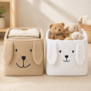Puede incluir: Dos cestas de almacenamiento cuadradas hechas de tela suave y texturizada. La cesta izquierda es de color tostado con un diseño de cara de perro, incluyendo orejas caídas y nariz y boca bordadas en negro. La cesta derecha es blanca con un diseño similar.