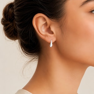 Puede incluir: Un pendiente pequeño de plata pulida con forma de gota alargada y lisa. El pendiente presenta una superficie metálica reflectante y un diseño sencillo que cuelga directamente del lóbulo de la oreja con una sujeción discreta y segura.