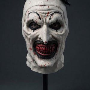 Op de afbeelding: Een clownmasker voor het hele hoofd met een bleke witte teint met rode bloedspatten, zwarte oogmake-up en een brede, donkere mond die rode tanden onthult. Het masker is voorzien van een kleine zwarte hoge hoed en staat op een zwarte standaard.
