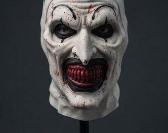 May include: A full-head clown mask featuring a pale white complexion with red blood splatters, black eye makeup, and a wide, dark mouth revealing red teeth. The mask is topped with a small black top hat and is displayed on a black stand.