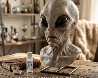 Puede incluir: Una máscara alienígena realista de piel gris, grandes ojos negros y rasgos definidos, montada en un soporte de metal. Junto a ella hay una pequeña botella de vidrio con la etiqueta Formula X-9, una brújula y un libro encuadernado en cuero.