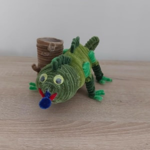 Peut inclure: Une figurine de lézard verte fabriquée à partir de cure-pipes enroulés, avec des yeux mobiles, une bouche rouge et une petite langue en pompon bleu. Le lézard est placé à côté d'un petit tube cylindrique marron en matière enroulée.