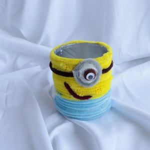Peut inclure: Un récipient cylindrique recouvert de cure-pipes duveteux jaunes et bleu clair. La partie supérieure présente un cadre oculaire circulaire gris avec un œil mobile blanc et noir, une fine bande horizontale brune et une bouche brune incurvée.