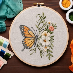 May include: A circular wooden embroidery hoop containing a light beige fabric panel. The fabric features a hand-stitched design of an orange and black butterfly positioned next to a cluster of white, pink, and yellow flowers with green leaves.