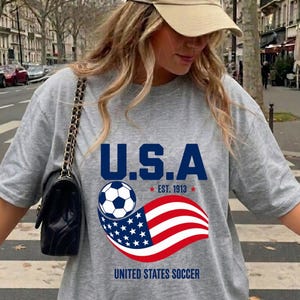 Może przedstawiać: Szary melanżowy t-shirt z kr&oacute;tkim rękawem z nadrukiem graficznym zawierającym napis U.S.A. oraz UNITED STATES SOCCER, piłkę nożną i stylizowaną amerykańską flagę z napisem EST. 1913, w zestawie z czarną torebką na ramię ze złotym łańcuszkiem.