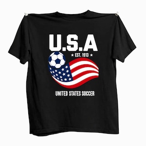 Może przedstawiać: Czarny t-shirt z kr&oacute;tkim rękawem z grafiką zawierającą napis U.S.A, EST. 1913 oraz UNITED STATES SOCCER. Wz&oacute;r przedstawia biało-czarną piłkę nożną nad stylizowaną, powiewającą amerykańską flagą w kolorach czerwonym, białym i niebieskim.