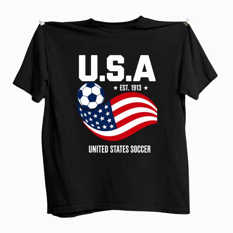 Może przedstawiać: Czarny t-shirt z kr&oacute;tkim rękawem z grafiką zawierającą napis U.S.A, EST. 1913 oraz UNITED STATES SOCCER. Wz&oacute;r przedstawia biało-czarną piłkę nożną nad stylizowaną, powiewającą amerykańską flagą w kolorach czerwonym, białym i niebieskim.