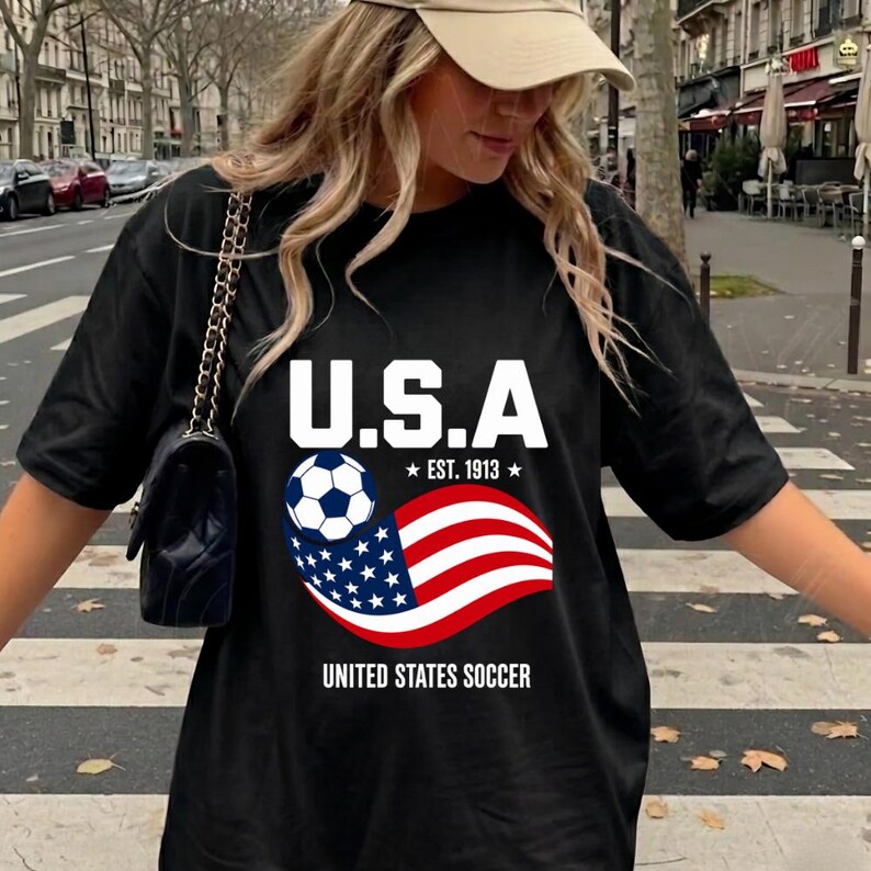 Może przedstawiać: Czarny t-shirt z nadrukiem graficznym z tekstem U.S.A. EST. 1913 oraz UNITED STATES SOCCER. Wz&oacute;r zawiera piłkę nożną i stylizowaną falę flagi amerykańskiej. Koszulka ma kr&oacute;tkie rękawy i okrągły dekolt, noszona z czarną torebką.