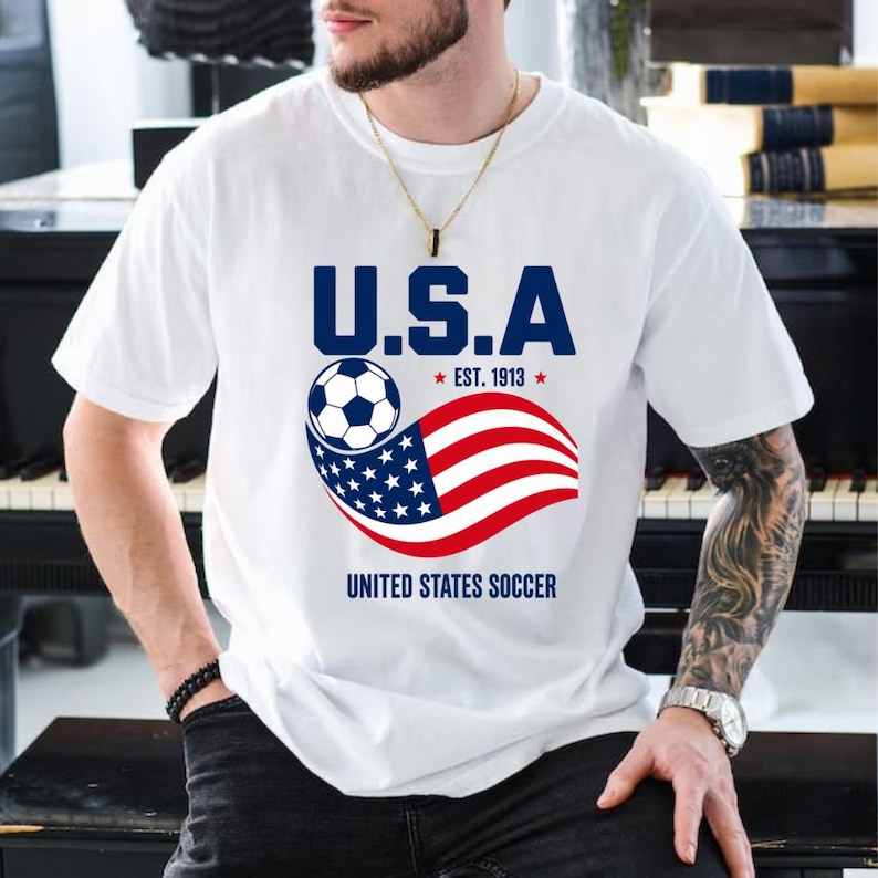 Może przedstawiać: Biały t-shirt z kr&oacute;tkim rękawem z grafiką zawierającą napis U.S.A. oraz EST. 1913 nad piłką nożną i falującą amerykańską flagą. Poniżej grafiki znajduje się napis UNITED STATES SOCCER wydrukowany pogrubionymi, niebieskimi wielkimi literami.