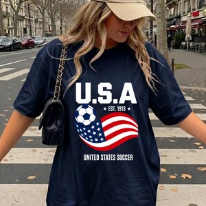 Może przedstawiać: Granatowy t-shirt z białą grafiką zawierającą napis U.S.A, EST. 1913 oraz UNITED STATES SOCCER. Wz&oacute;r przedstawia czarno-białą piłkę nożną oraz stylizowaną amerykańską flagę z czerwonymi, białymi i niebieskimi pasami oraz białymi gwiazdami.
