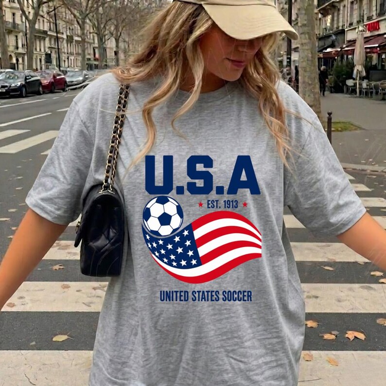 Może przedstawiać: Szary melanżowy t-shirt z kr&oacute;tkim rękawem z nadrukiem graficznym zawierającym napis U.S.A. oraz UNITED STATES SOCCER, piłkę nożną i stylizowaną amerykańską flagę z napisem EST. 1913, w zestawie z czarną torebką na ramię ze złotym łańcuszkiem.