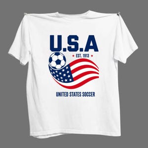 Może przedstawiać: Biały t-shirt z kr&oacute;tkim rękawem z grafiką zawierającą napis U.S.A, EST. 1913 oraz UNITED STATES SOCCER. Wz&oacute;r przedstawia czarno-białą piłkę nożną nad stylizowaną, falującą amerykańską flagą w kolorach czerwonym, białym i niebieskim.