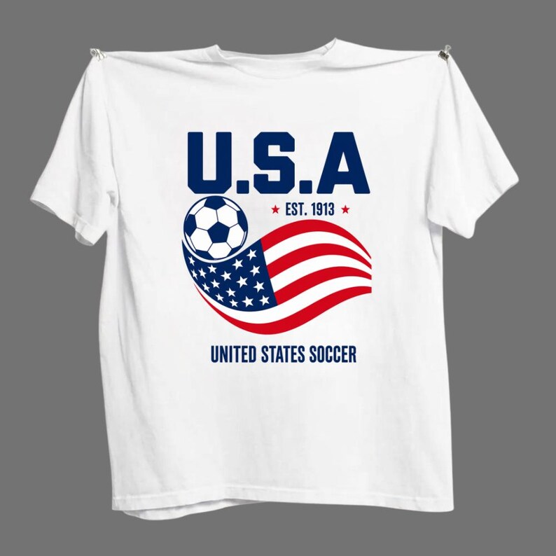 Może przedstawiać: Biały t-shirt z kr&oacute;tkim rękawem z grafiką zawierającą napis U.S.A, EST. 1913 oraz UNITED STATES SOCCER. Wz&oacute;r przedstawia czarno-białą piłkę nożną nad stylizowaną, falującą amerykańską flagą w kolorach czerwonym, białym i niebieskim.