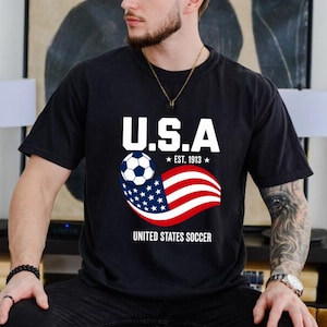 Może przedstawiać: Czarny t-shirt z kr&oacute;tkim rękawem z grafiką na piersi, zawierającą napis U.S.A. oraz UNITED STATES SOCCER, piłkę nożną i falującą amerykańską flagę z napisem EST. 1913 w kolorach białym, czerwonym i niebieskim.