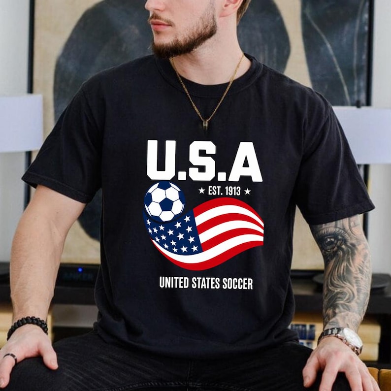 Może przedstawiać: Czarny t-shirt z kr&oacute;tkim rękawem z grafiką na piersi, zawierającą napis U.S.A. oraz UNITED STATES SOCCER, piłkę nożną i falującą amerykańską flagę z napisem EST. 1913 w kolorach białym, czerwonym i niebieskim.