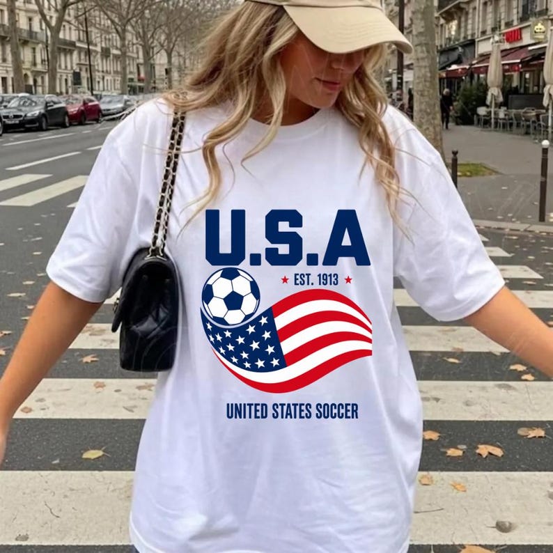 Może przedstawiać: Biały t-shirt z kr&oacute;tkim rękawem z nadrukiem graficznym zawierającym tekst U.S.A. EST. 1913 oraz UNITED STATES SOCCER. Wz&oacute;r przedstawia piłkę nożną oraz stylizowaną falę amerykańskiej flagi w kolorach czerwonym, białym i niebieskim na środku.