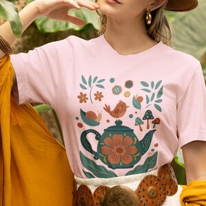Pode incluir: Uma t-shirt de manga curta rosa claro com uma estampa gráfica de um bule verde-azulado escuro com uma flor laranja grande, um pequeno pássaro castanho, um caracol e cogumelos, combinada com uma saia branca e um cinto castanho circular.