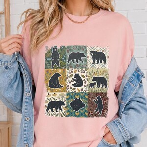 Puede incluir: Una camiseta de cuello redondo rosa claro con una cuadrícula de nueve ilustraciones cuadradas. Cada cuadrado muestra la silueta de un oso negro sobre diversos patrones botánicos, florales y forestales en tonos verdes, amarillos y marrones.