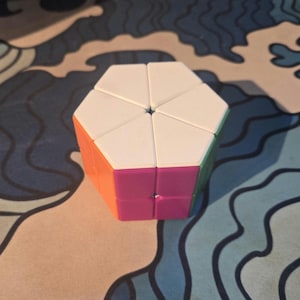 Könnte beinhalten: Ein hexagonales Puzzle-Spielzeug mit einer Oberseite, die in sechs weiße dreieckige Segmente unterteilt ist. Die Seitenflächen zeigen leuchtende Farben wie Orange, Magenta und Grün. Das Objekt hat eine glatte Kunststoffoberfläche.