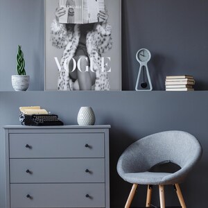 May include: A framed black and white wall art print featuring a person holding a Vogue magazine in front of their face, wearing a patterned coat. The print displays the word VOGUE in large white letters and is displayed on a dark gray wall.