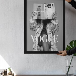 May include: A black and white framed wall art print featuring a person wearing a leopard print coat, holding an open Vogue magazine in front of their face. The magazine displays the text Vogue and Balenciaga. The frame is thin and black.