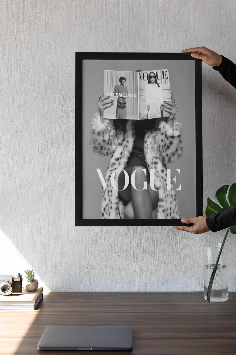 May include: A black and white framed wall art print featuring a person wearing a leopard print coat, holding an open Vogue magazine in front of their face. The magazine displays the text Vogue and Balenciaga. The frame is thin and black.
