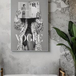 May include: A framed black and white wall art print featuring a person wearing a spotted faux fur coat, holding an open Vogue magazine in front of their face. The magazine displays the text Balenciaga and Vogue. The word VOGUE is printed in large white letters across the lower portion of the artwork.