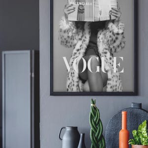 May include: A black and white framed wall art print featuring a person wearing a leopard print coat, holding a Vogue magazine open in front of their face. The magazine displays the text Balenciaga and Vogue. The word VOGUE is printed in large white letters across the center of the artwork.