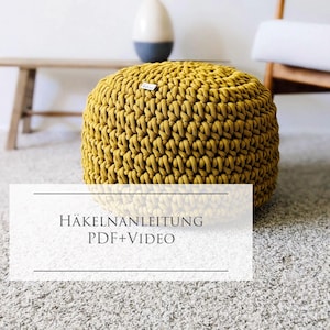 May include: A round, mustard yellow crocheted pouf with a thick, textured yarn construction and a small white brand label on the side. The image includes the text HÄKELANLEITUNG PDF+VIDEO overlaid on a white rectangular background at the bottom.