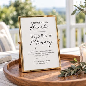 May include: A vertical gold metal frame holding a white sign with black text that reads: A MOMENT TO REMEMBER SHARE A MEMORY. Please take a moment to share a story, a memory, or a kind word about our loved one. Your memories mean so much to us.