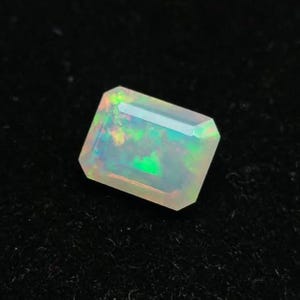 May include: A rectangular faceted opal gemstone featuring a translucent body with vibrant flashes of green, yellow, and orange play-of-color. The stone has a polished surface with sharp edges and a clear, multi-faceted cut that reflects light.