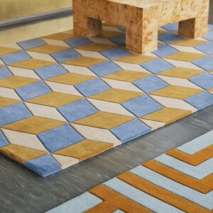 以下が含まれることがあります： 青、マスタードイエロー、クリーム色の菱形が繰り返される幾何学模様の長方形のラグ。手前には、マスタードイエローとライトブルーの迷路のような直線的な模様のラグが一部見えています。