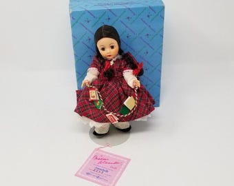 Może przedstawiać: Lalka z ciemnymi włosami ubrana w czerwono-zieloną sukienkę w kratę z białą koronką, trzymająca sznurek z miniaturowymi książkami. Lalka stoi przed niebieskim pudełkiem we wzory oraz różową metką z napisem Madame Alexander Annabelle 2253.