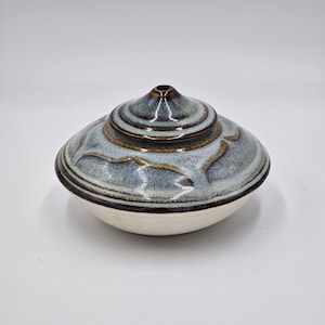 May include: A ceramic lidded bowl featuring a rounded base with a smooth off-white finish. The lid is dome-shaped with a central knob, decorated in a mottled blue and grey glaze with dark brown accents along the edges and a petal-like pattern.