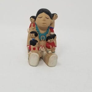 May include: A ceramic figurine depicting a seated figure with multiple smaller figures. The main figure has black hair, a turquoise necklace, and is wearing a tan dress with red accents. The smaller figures are wearing red and black clothing.
