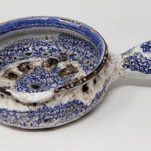 May include: A ceramic bowl with a handle, featuring a speckled blue and white design. The rim is a darker blue, and the handle has a rounded shape. The bowl has a rustic, handmade appearance.