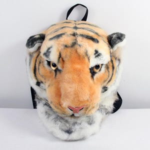 May include: A plush tiger head backpack featuring realistic orange, black, and white fur patterns. The design includes pointed ears, amber-coloured eyes, a pink nose, and long white whiskers. A black fabric carrying handle is attached to the top.