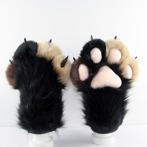 May include: A pair of plush animal paw gloves featuring long black and tan faux fur. One glove shows the back with sharp black claws, while the other displays the palm side with soft, light pink padded paw pads and matching black claws.