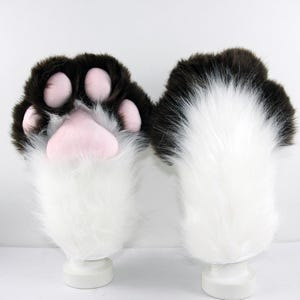May include: A pair of plush animal paw gloves featuring long white faux fur on the cuffs and dark brown faux fur on the upper hand. The palm side displays four soft pink rounded toe pads and one large pink central pad, mimicking an animal paw.
