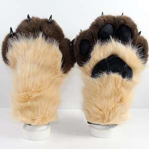 May include: A pair of plush animal paw gloves featuring long tan faux fur on the palms and dark brown faux fur on the back. The gloves include black padded paw pads and sharp, pointed black claws extending from the tips of each finger.