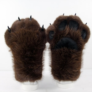 May include: A pair of brown faux fur gloves designed to resemble animal paws. One glove displays four black pointed claws on the top, while the other shows a black padded paw print on the palm side. The brown fur is long, textured, and covers the entire glove.
